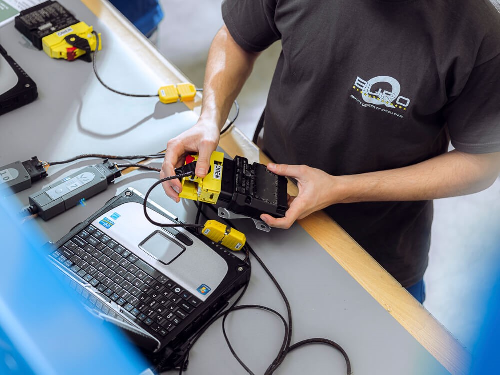 A person wearing a EUROQ T-shirt is working on an electronic vehicle component that is connected to several cables and a laptop. The scene depicts the diagnosis or programming of automotive parts using specialized software and measuring devices as part of quality and function testing.