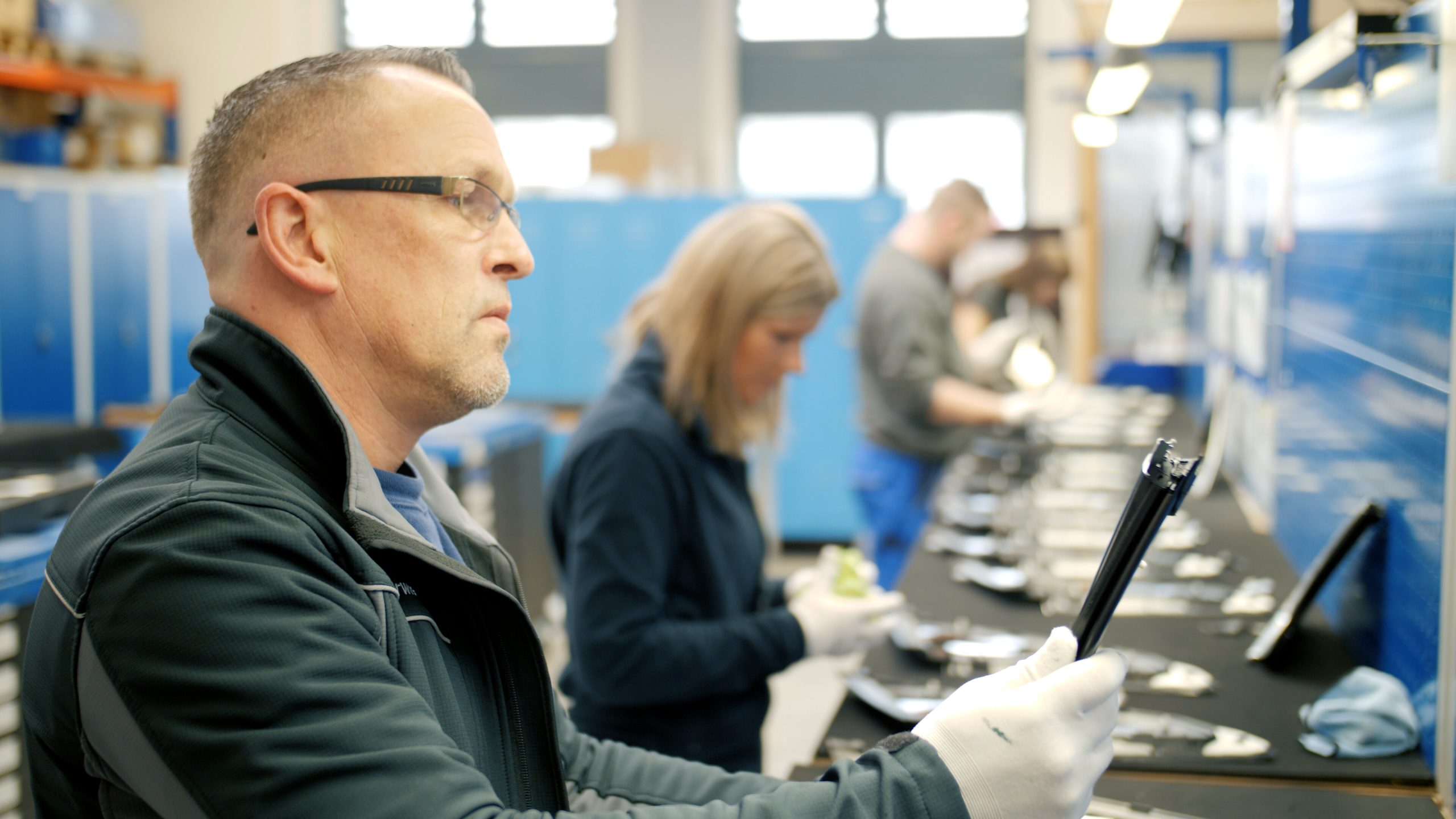 Ein Mitarbeiter mit Schutzbrille und Handschuhen prüft ein Fahrzeugbauteil an einem langen Arbeitstisch. Im Hintergrund kontrollieren weitere Mitarbeitende Bauteile in einer Produktionshalle mit blauen Werkzeugwänden. Die Szene steht für Qualitätskontrolle, Kommissionieren und Umpacken in der Automobilindustrie.