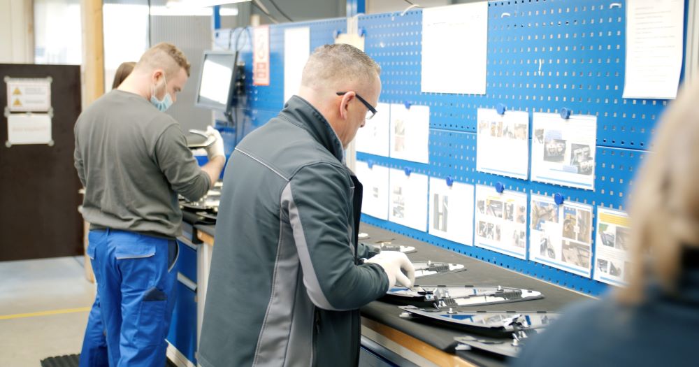 Several employees in workwear are performing quality control and assembly tasks on vehicle components at a workbench. In the background, a blue tool wall and information sheets are visible.